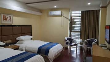 A hotel room with two beds, a desk, a chair, an air conditioning unit, and a window with blinds.