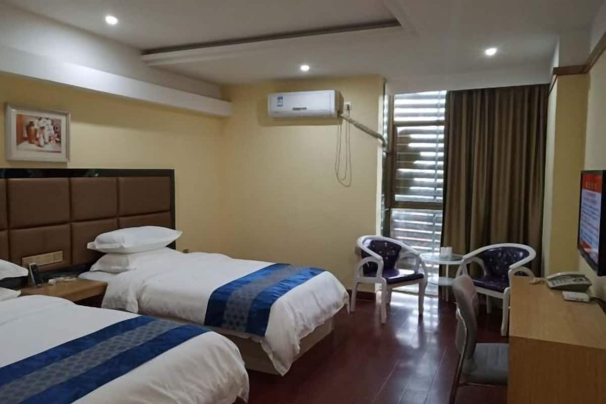 A hotel room with two beds, a desk, a chair, an air conditioning unit, and a window with blinds.