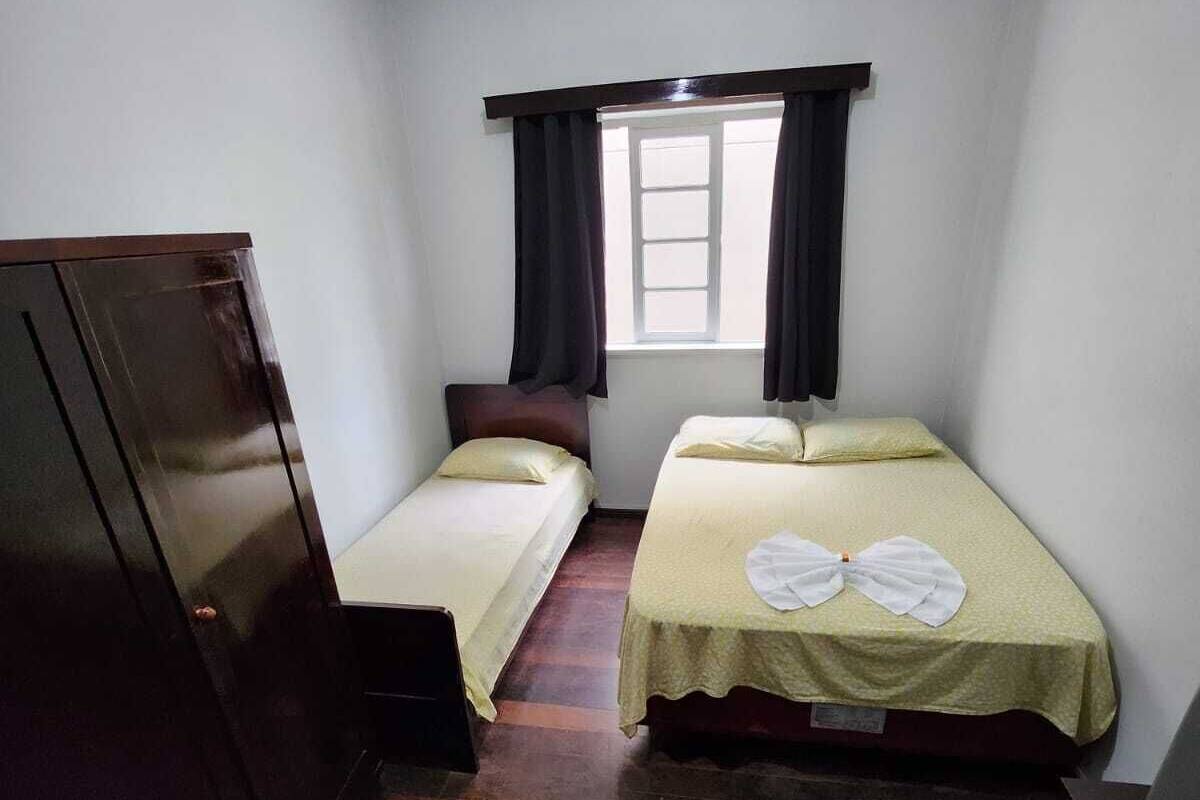 Um quarto pequeno e pouco iluminado com duas camas de solteiro, um guarda-roupa e uma janela com cortinas escuras.