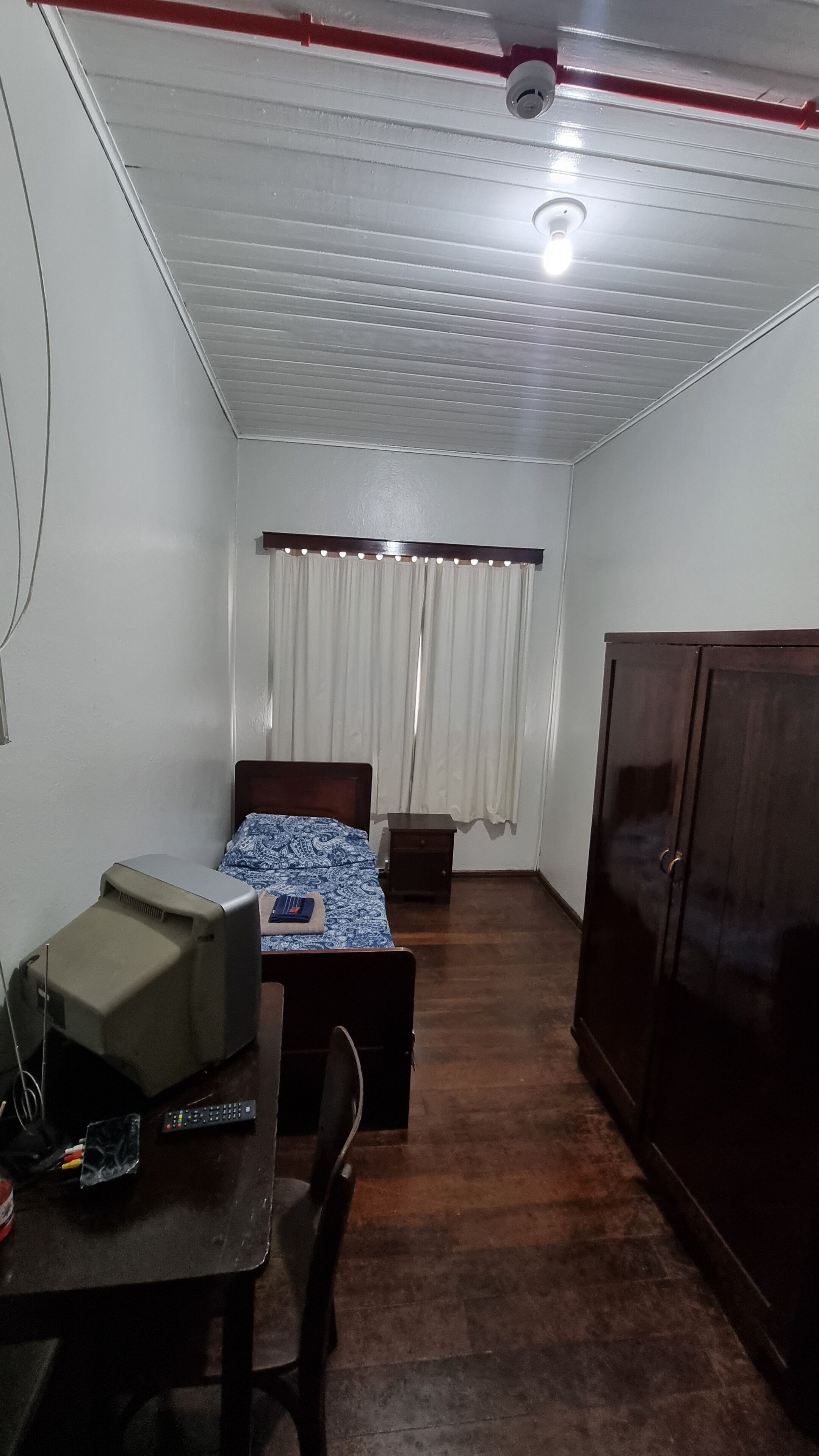 A small, dimly lit room with a single bed, a desk with a computer and a printer, a wardrobe, and a window with curtains.