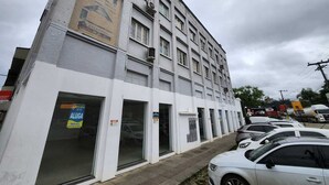 A multi-story building with a white facade, several ground-level commercial spaces, and a row of parked cars in front. - Hotel Real Koerich 24h (Rio do Sul)