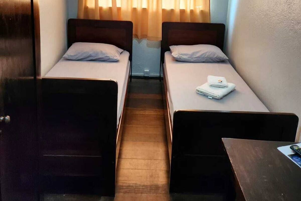 Quarto pequeno e compacto com duas camas de solteiro, guarda-roupa de madeira, uma mesinha e janela com cortinas.