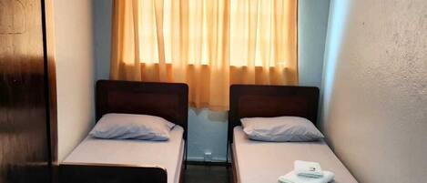 Una habitación pequeña y funcional con dos camas individuales, un armario de madera, una mesita y una ventana con cortinas.