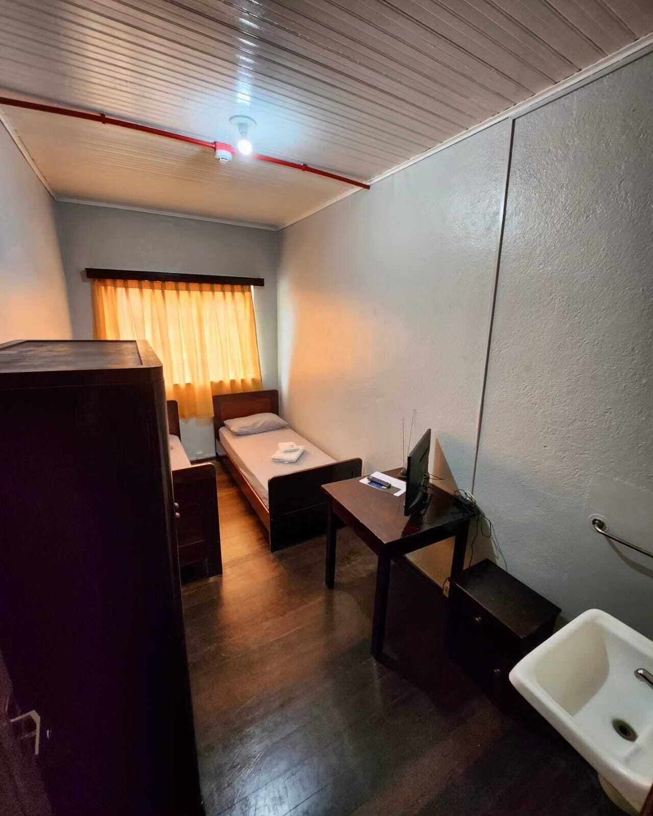 A compact hotel room with a single bed, a desk with a computer, a small sink, and a window with curtains.