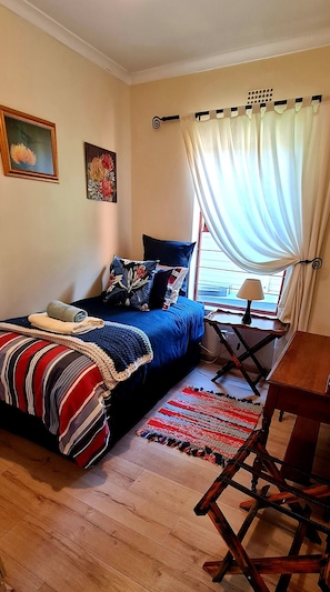 Quarto com cama, escrivaninha, cadeira, janela com cortinas e obras de arte nas paredes.