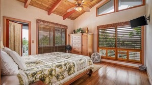 A bedroom with a wooden ceiling, a bed with a floral bedspread, a dresser, a TV mounted on the wall, and large windows with blinds. - Walk to Beach! HUGE 5 bedroom, 5 bath with Pool!! (Kailua-Kona)