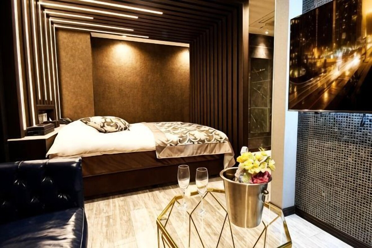 Une chambre d’hôtel moderne comprenant un lit, une table de chevet, un fauteuil en cuir, un vase décoratif avec des fleurs et un téléviseur fixé au mur.