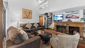 Un salon moderne avec un canapé en cuir marron, une table basse et une cheminée. Un escalier mène à un niveau supérieur, et une grande fenêtre offre une vue sur les montagnes.