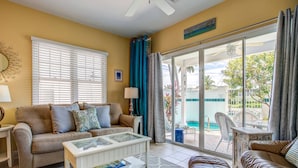 Un salon avec un canapé beige, une table basse blanche et une vue sur une piscine, visible à travers de grandes portes vitrées.