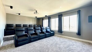 A home theater room with a carpeted floor, a ceiling fan, and a large window with curtains.