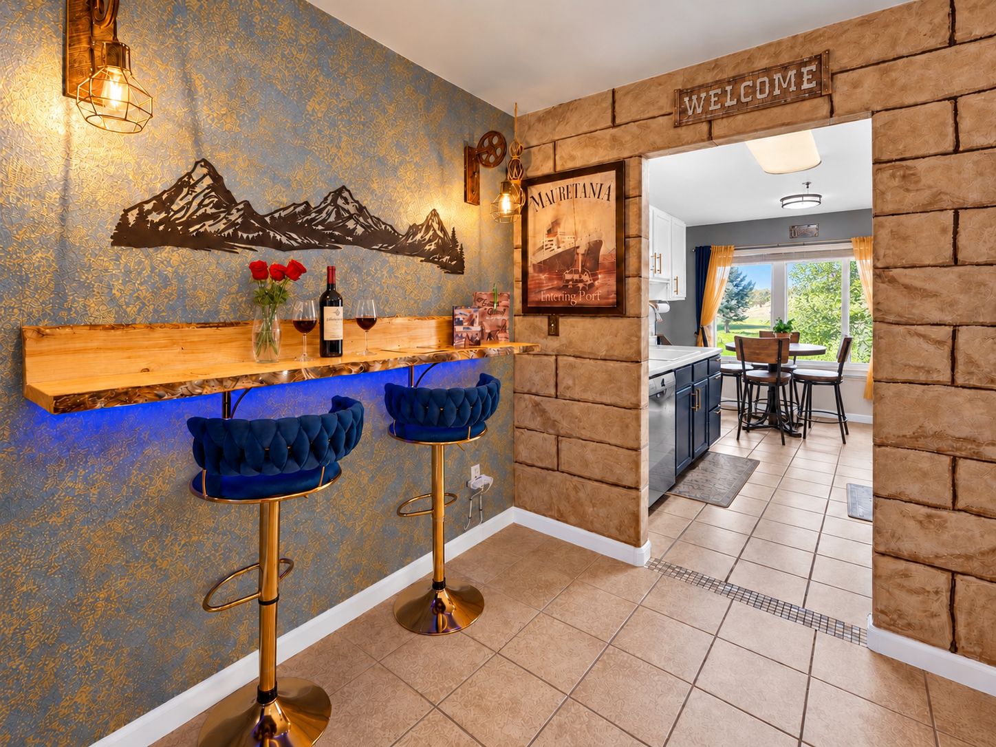 A bar area with a wooden shelf, two blue bar stools, a wine bottle, and glasses on the counter.