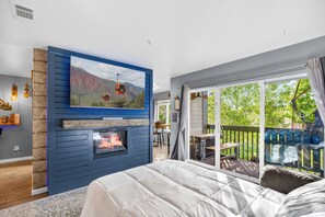 Een slaapkamer met een open haard, een bed, een televisie bevestigd aan een blauwe muur en een balkon met uitzicht op groen.