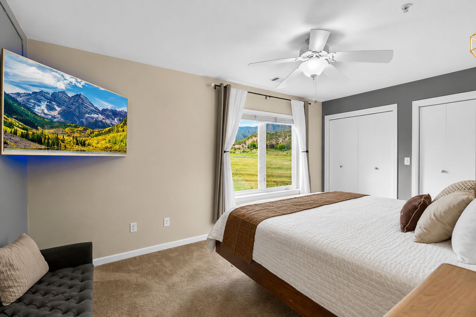 A bedroom with a bed, a ceiling fan, a large window with curtains, a painting of mountains, and a view of a green landscape.