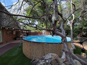 A backyard with a swimming pool surrounded by a wooden deck, a tree, and a brick house.