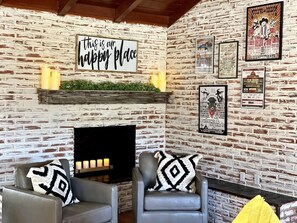 A cozy living room with a brick wall, a fireplace, two armchairs with patterned cushions, and a mounted sign above the fireplace.