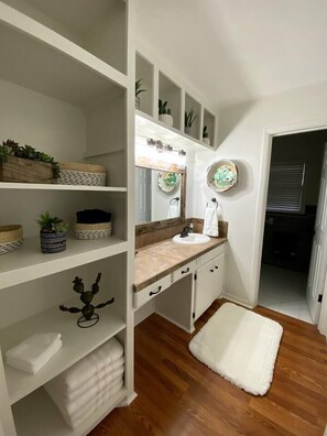 A bathroom with a white vanity, a sink, a mirror, and shelves with plants and baskets.