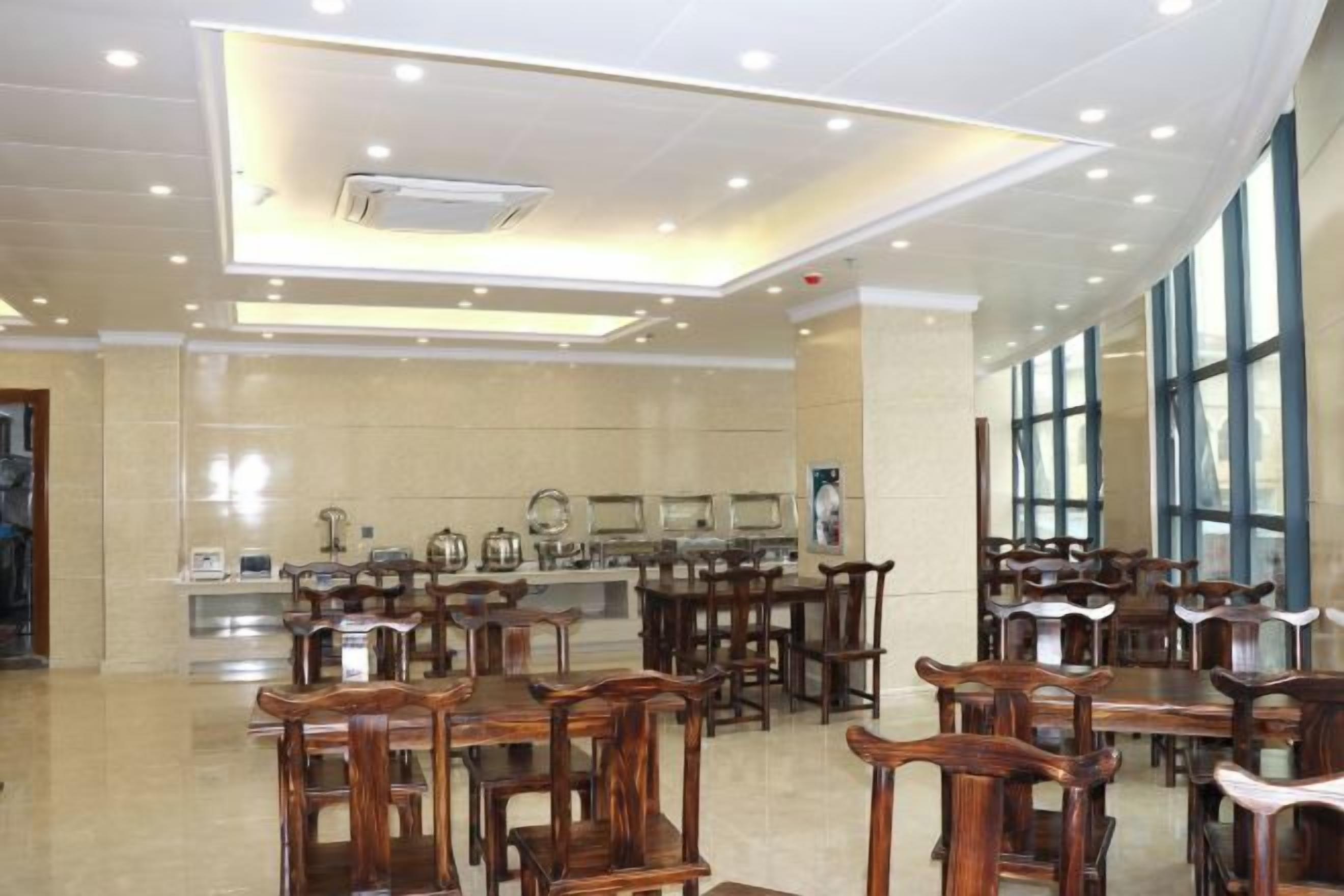 A spacious dining area with multiple wooden tables and chairs, a kitchen area in the background, and large windows allowing natural light.