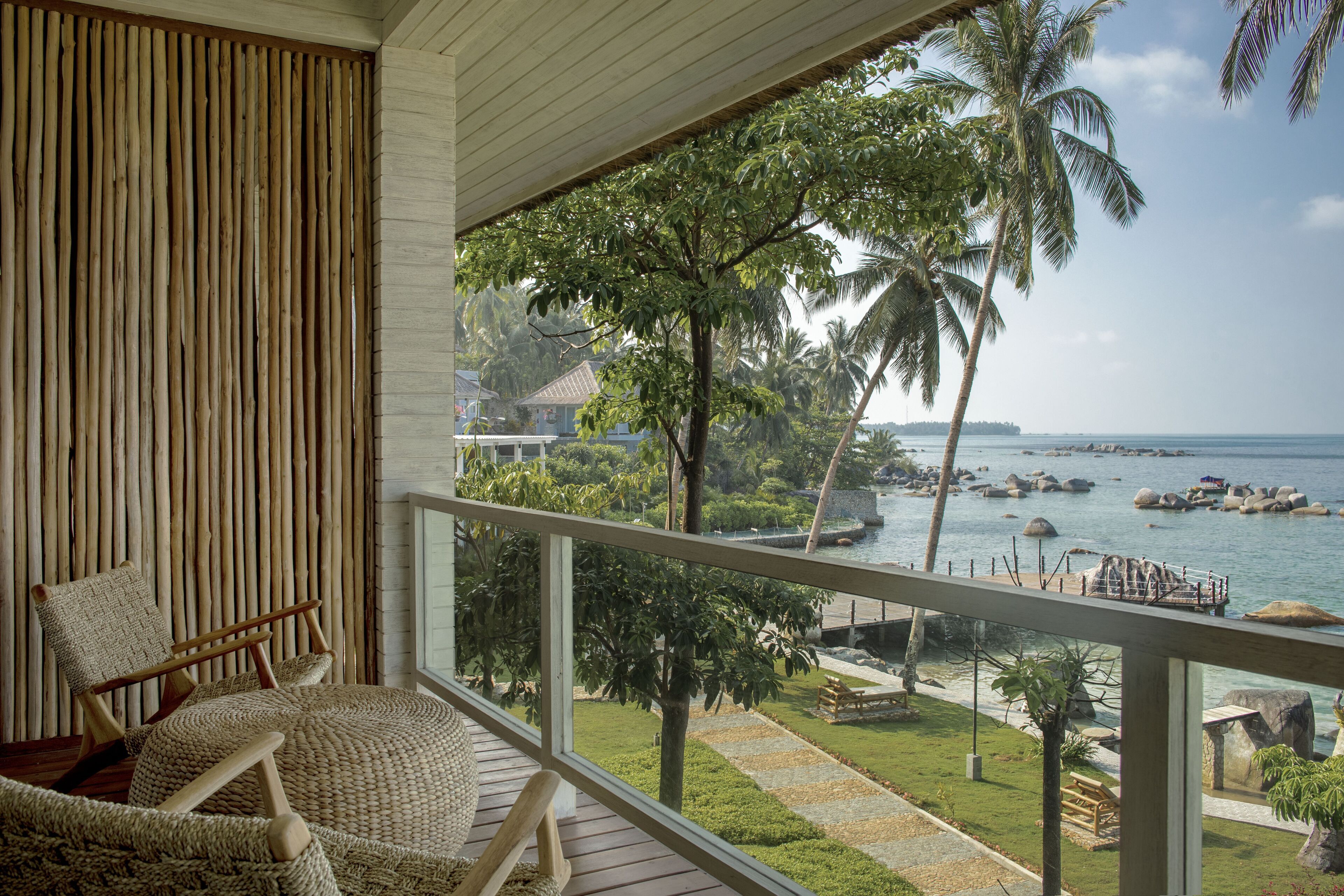 A balcony with a wicker chair, a view of a tropical landscape with palm trees and a body of water.
