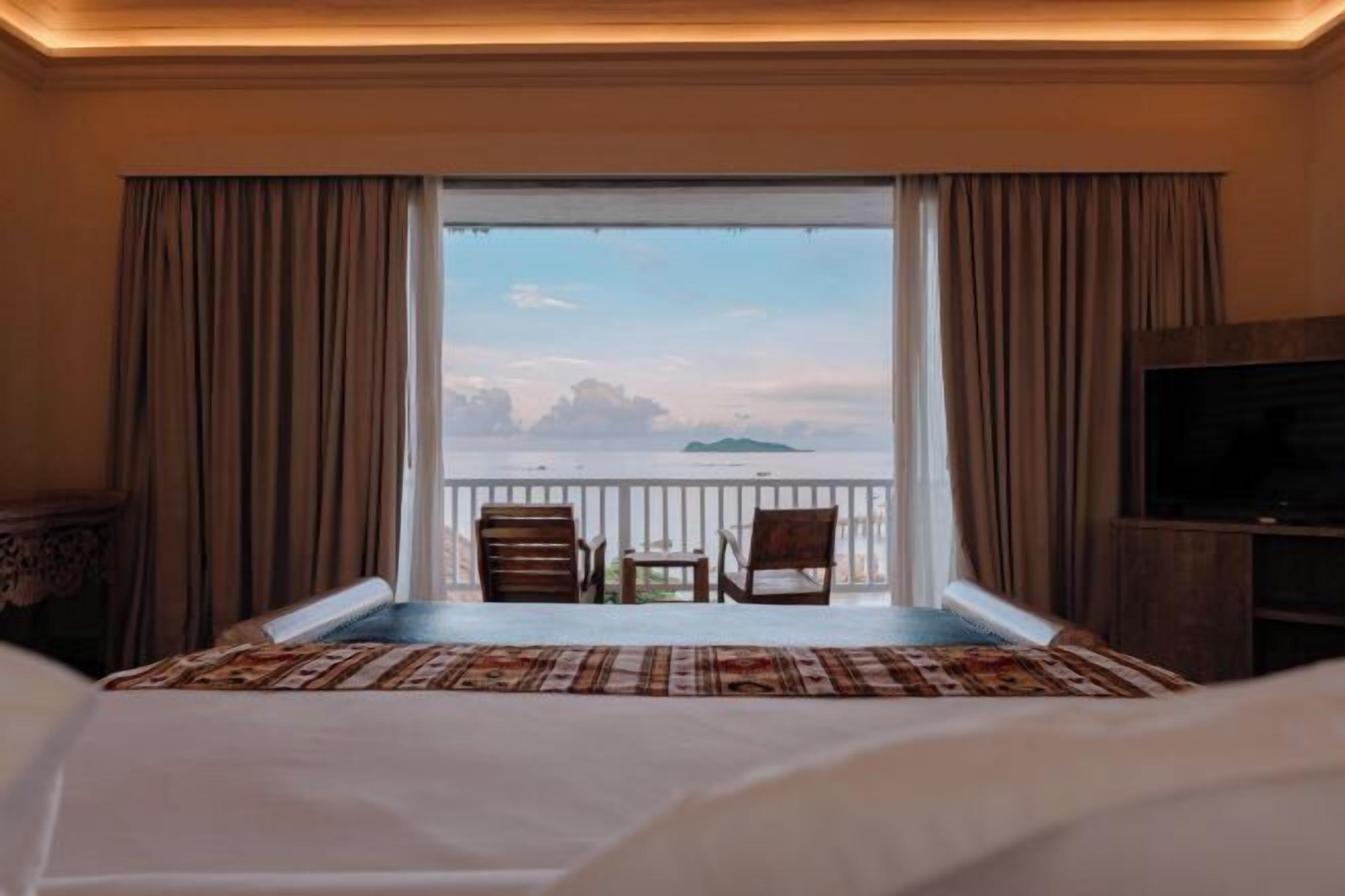 A hotel room with a bed, a TV, a balcony with a view of the sea, and two chairs.