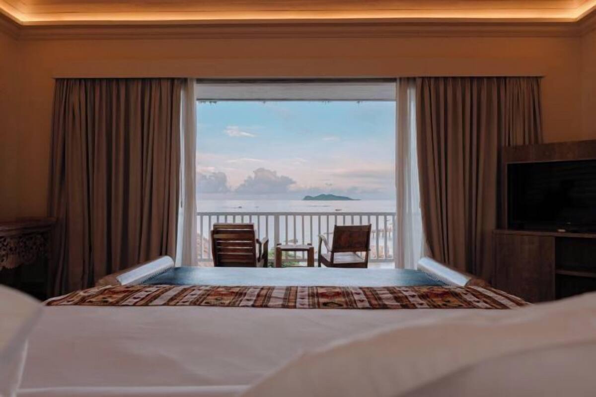 Habitación de hotel con cama, televisión, balcón y vista al mar, además de dos sillas.