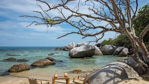 A beach with clear water, large rocks, and a wooden table with chairs.