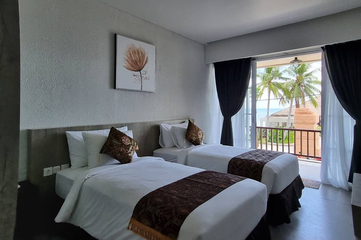 Habitación de hotel con dos camas, un ventanal con cortinas, vistas a palmeras y al mar, y una obra de arte enmarcada en la pared.