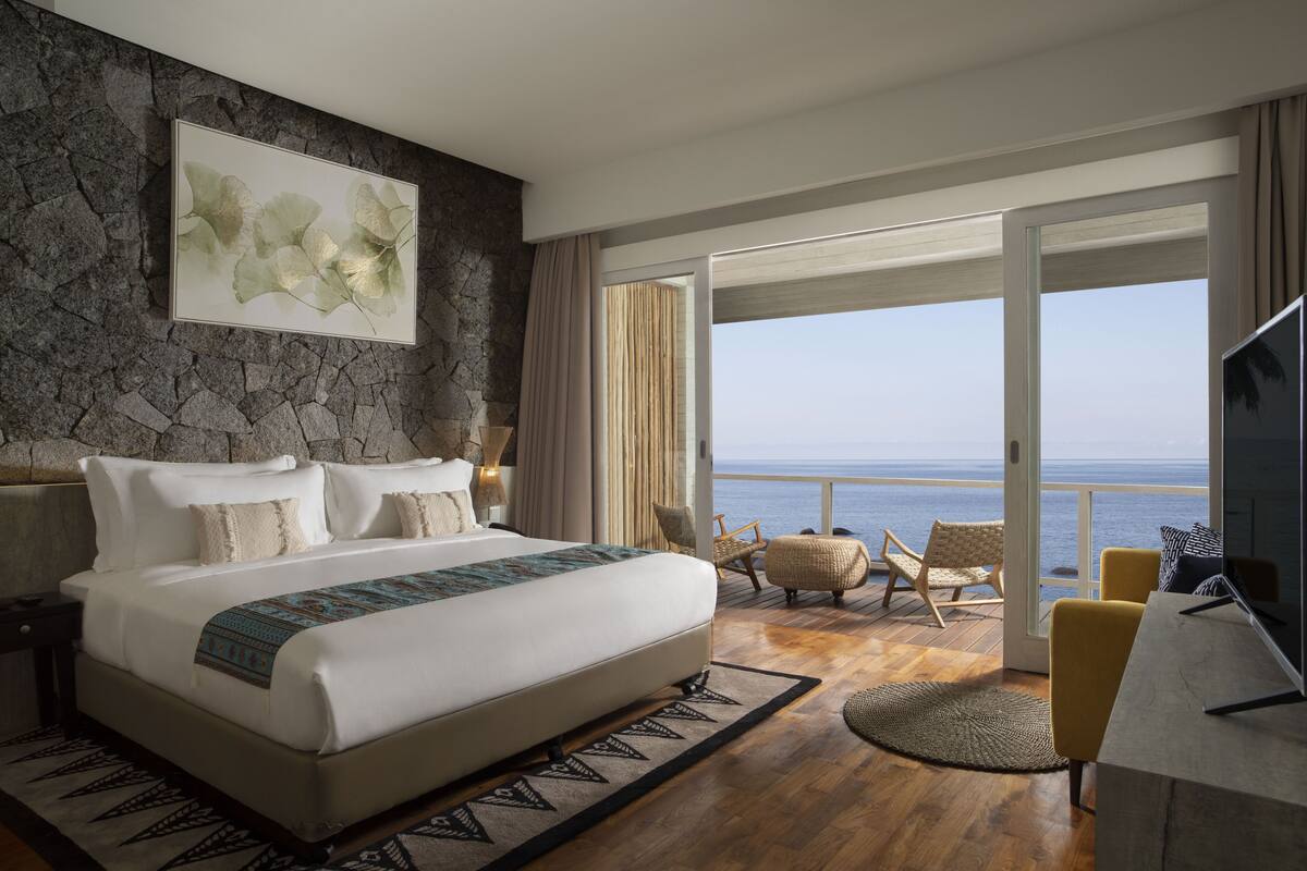 Un dormitorio amplio con una cama grande, una pared de acento de piedra, un balcón con vistas al mar y un televisor sobre una mesa de madera.