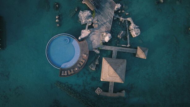 An aerial view of a resort with a central swimming pool, multiple overwater bungalows, and a connecting bridge.
