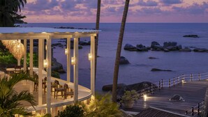 A beachfront restaurant with a wooden deck, tables, and chairs, overlooking the ocean and palm trees.