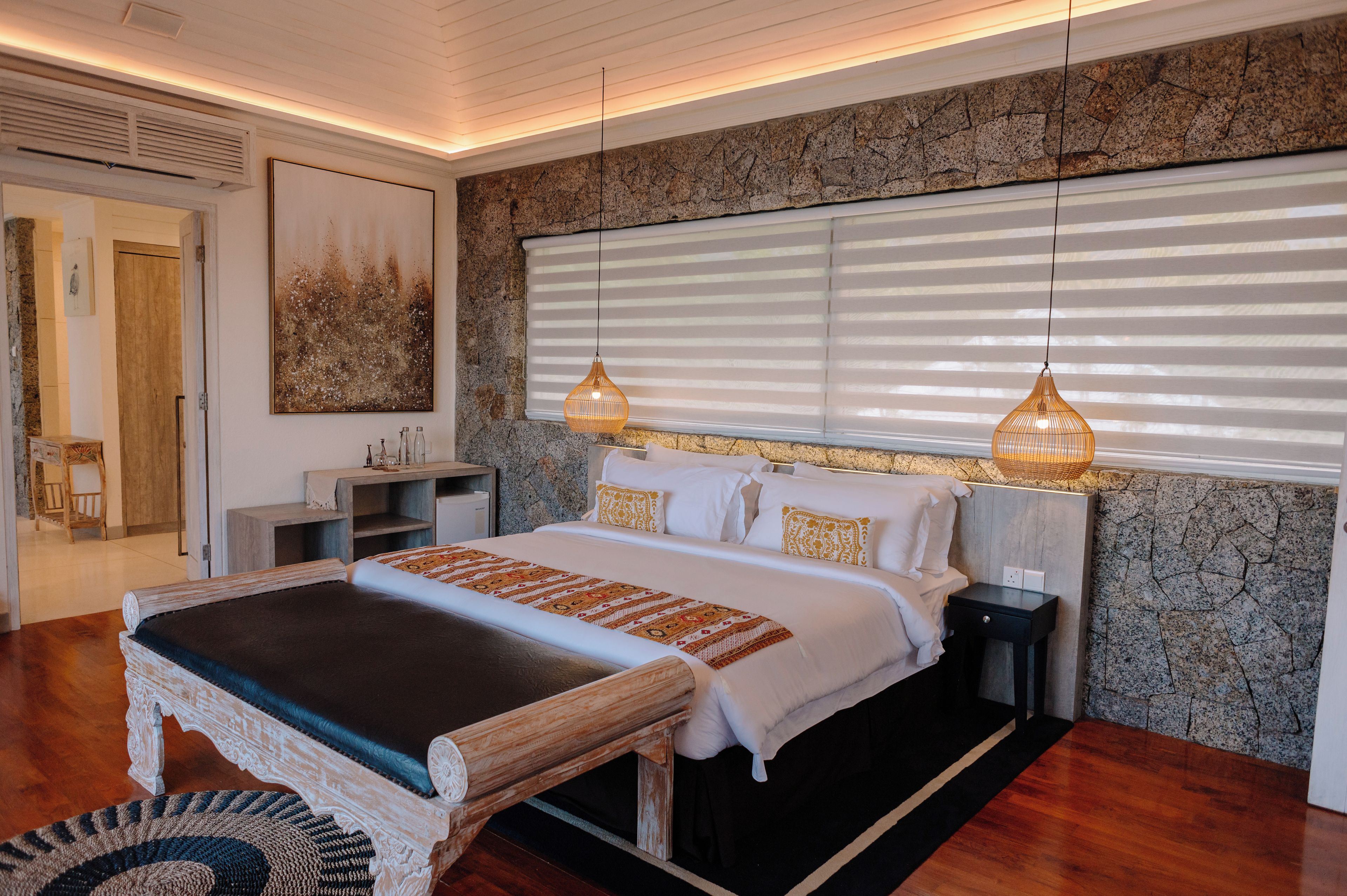 A bedroom with a large bed, a nightstand, a bedside table, a wall-mounted artwork, and a window with blinds.