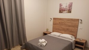 Un lit bien fait, recouvert d’une courtepointe grise matelassée, avec deux serviettes enroulées posées sur le pied de lit. On y trouve une tête de lit en bois, une petite table de chevet avec une lampe, et un tableau accroché au mur au-dessus du lit.