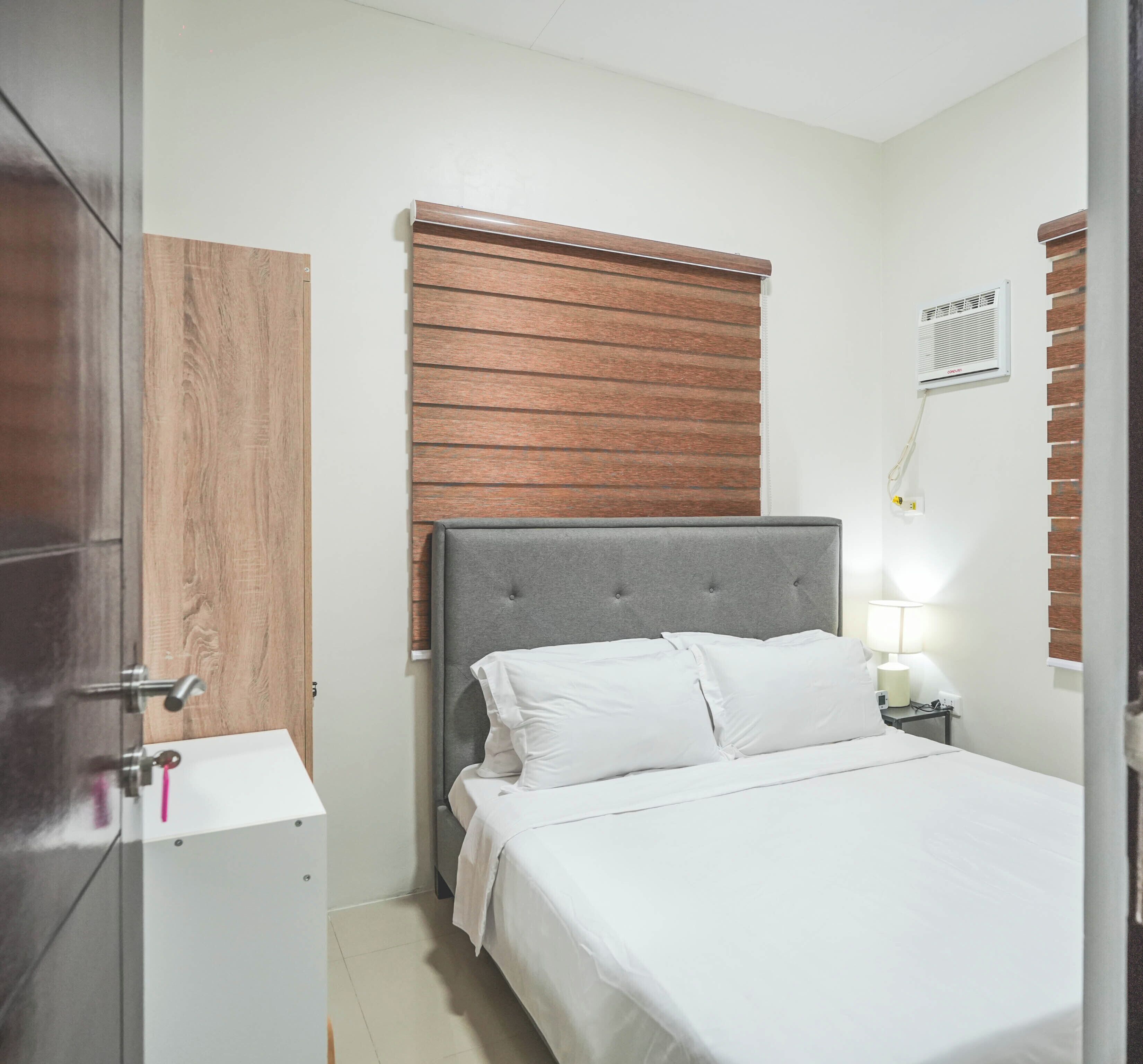 Une chambre à coucher comprenant un lit, une table de chevet, un climatiseur fixé au mur et une porte en bois.