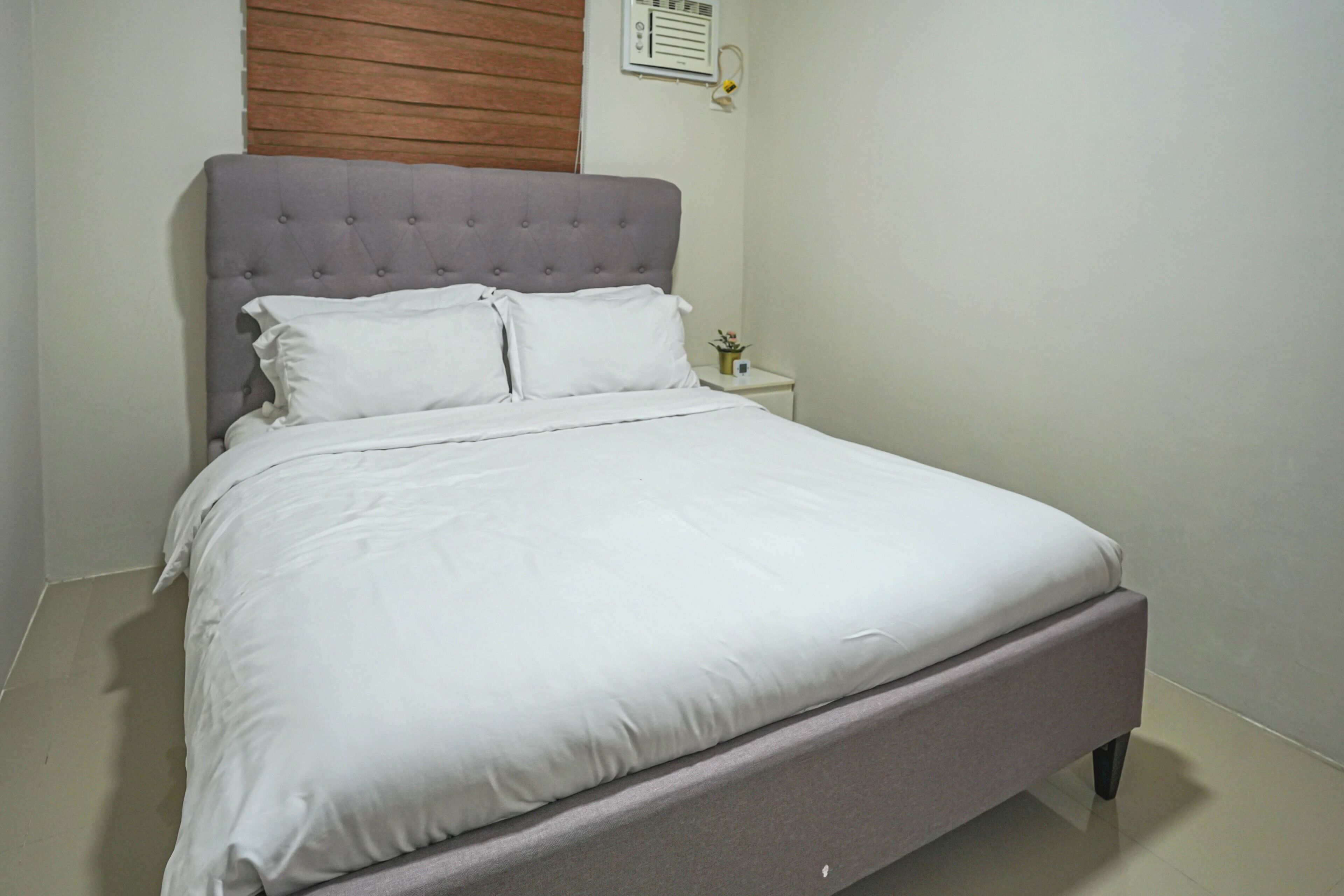 Cama bem arrumada com cabeceira cinza e roupa de cama branca em quarto com parede de painel de madeira e uma pequena mesa de cabeceira com uma planta.