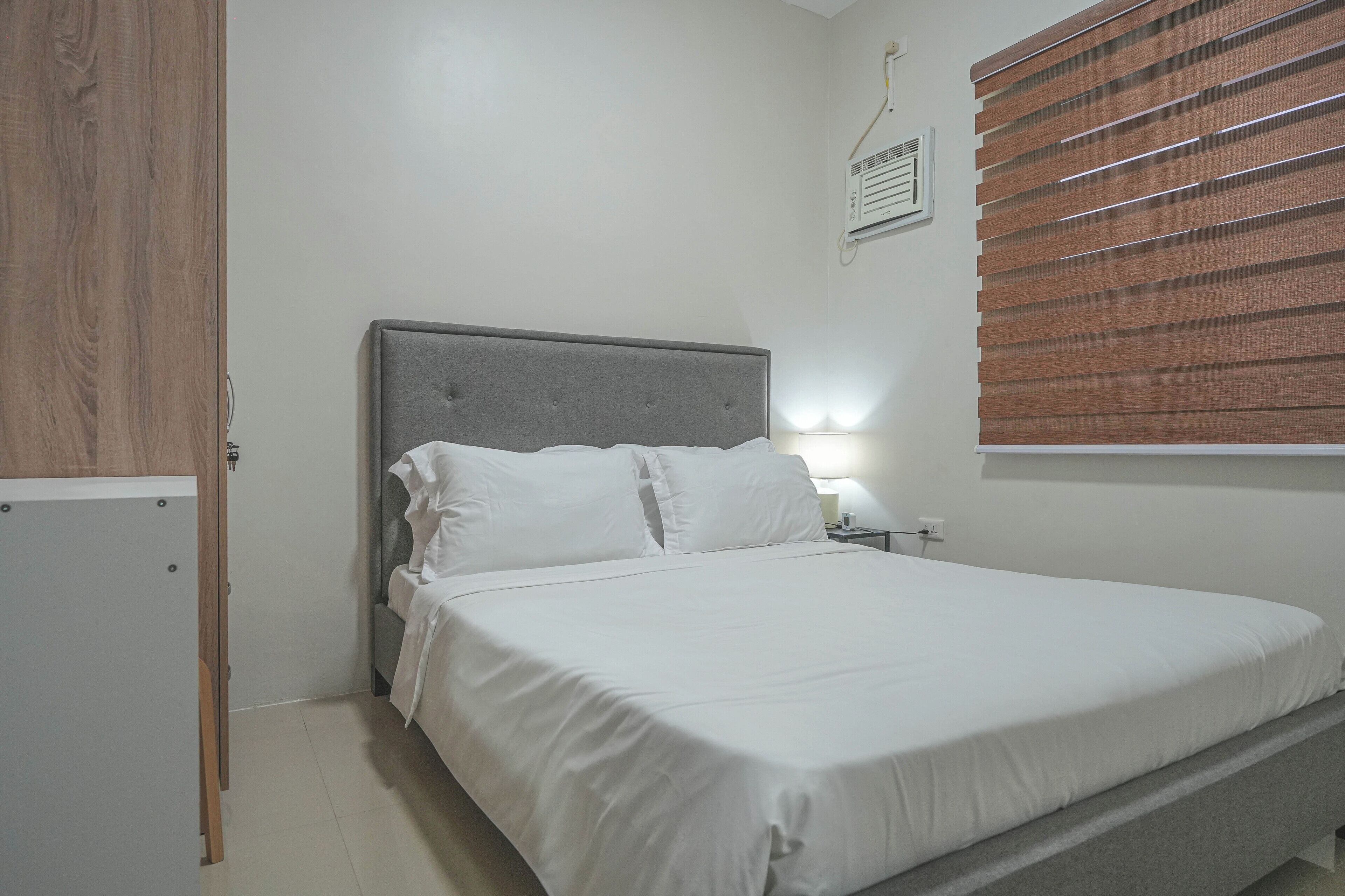 Een slaapkamer met een bed, een nachtkastje, een wandheater en een houten verstelbaar raambekleden.