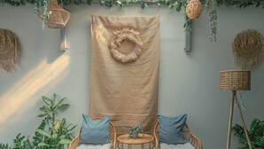 Una habitación con un gran tapiz tejido, sillas de mimbre con cojines y plantas en macetas.