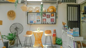 Une pièce équipée d'un ventilateur, de chaises en osier, d'une petite table avec une lampe et d'un présentoir mural où sont exposés divers objets.