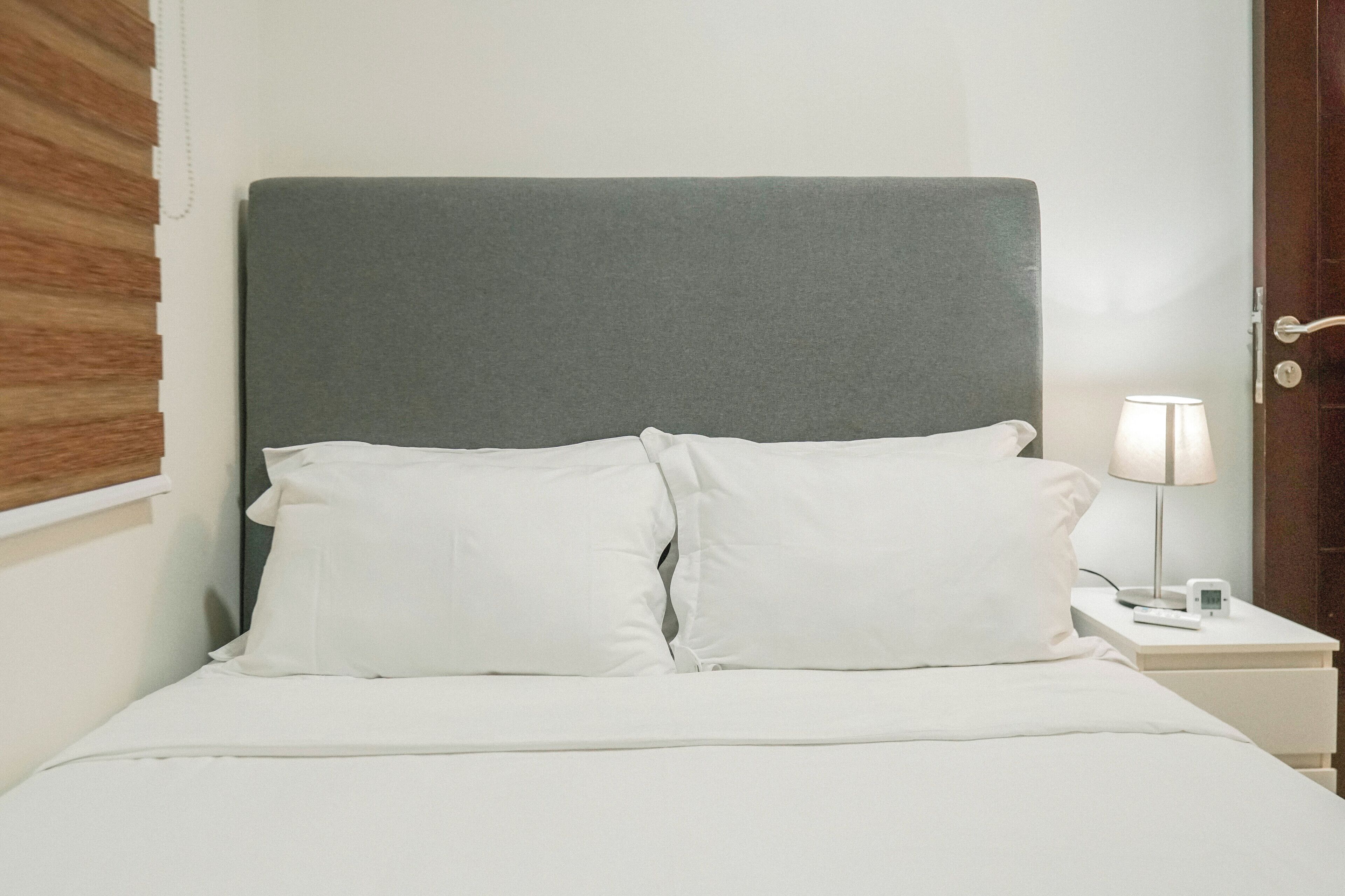 Una cama con cabecera gris, ropa de cama blanca y una mesita de noche con una lámpara y un reloj.