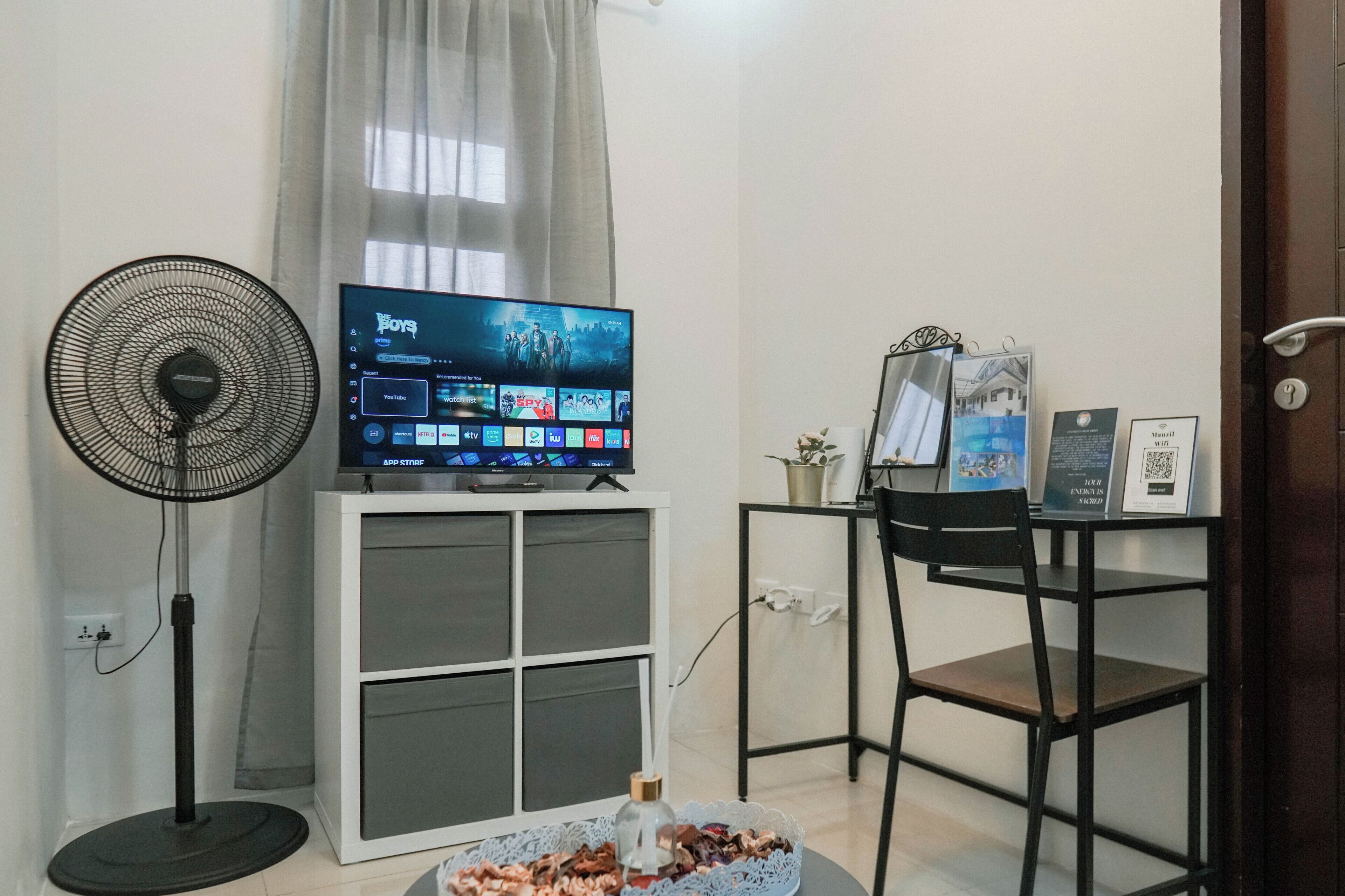 Een kamer met een televisie op een standaard, een ventilator, een bureau met een computer, en een deur met een QR-code.