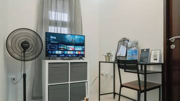 Een kamer met een televisie op een standaard, een ventilator, een bureau met een computer, en een deur met een QR-code.