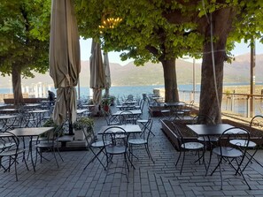 Um café ao ar livre com mesas, cadeiras, guarda-sóis e vista para a água e montanhas.