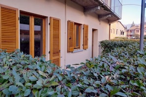 A building with wooden shutters, a concrete balcony, and dense green foliage in front.