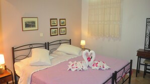 Un letto rifatto con cura, con una coperta rosa, cuscini bianchi e un originale accostamento di asciugamani a forma di cuore. In parete, quattro quadri incorniciati, una lampada sul comodino e una finestra con tenda leggera.