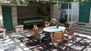 Une terrasse extérieure avec une table ronde et des chaises en bois, entourée de murs en pierre et d’une porte verte.