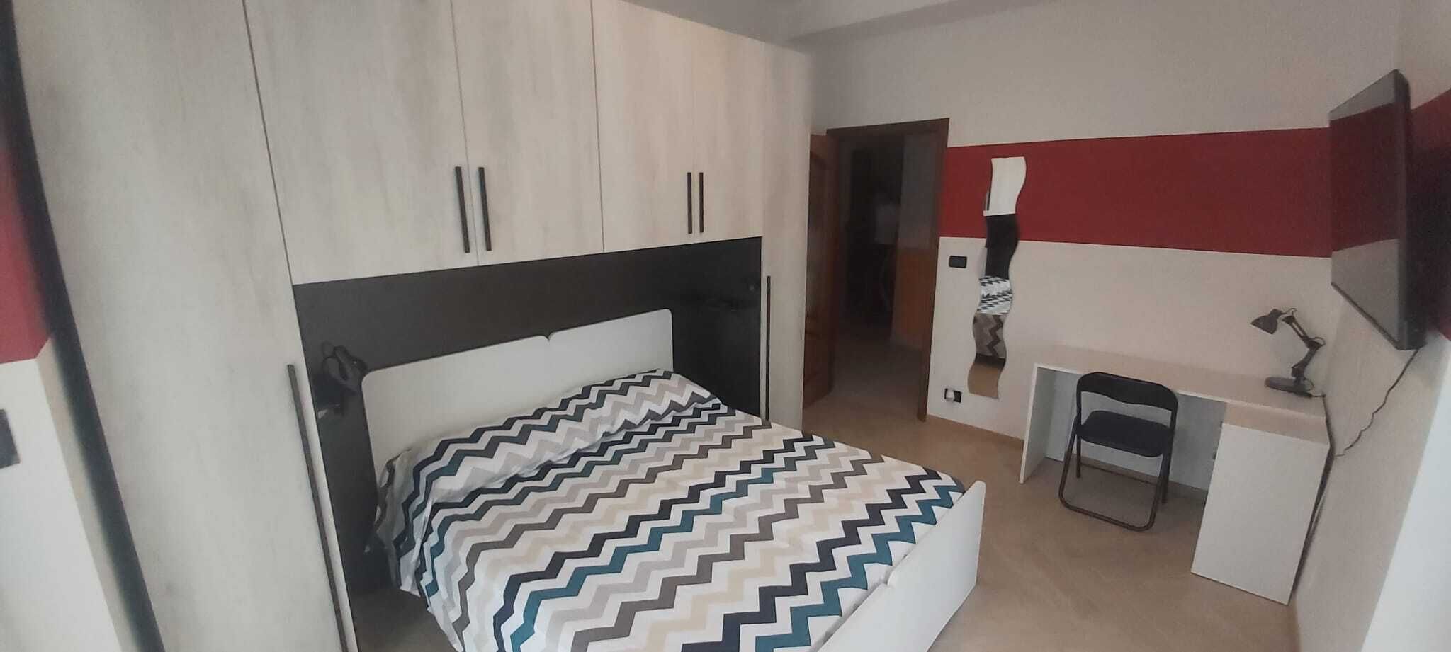 Ein Schlafzimmer mit Bett, Schreibtisch und Stuhl. Ein Kleiderschrank mit zwei Türen und ein Spiegel befinden sich über dem Bett.