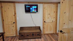 A room with a wooden chest, a mounted TV displaying streaming services, and wooden doors. - Cabin on a Secluded Cove -- READ DESCRIPTION REGARDING SAM RAYBURN WATER LEVEL (Broaddus)