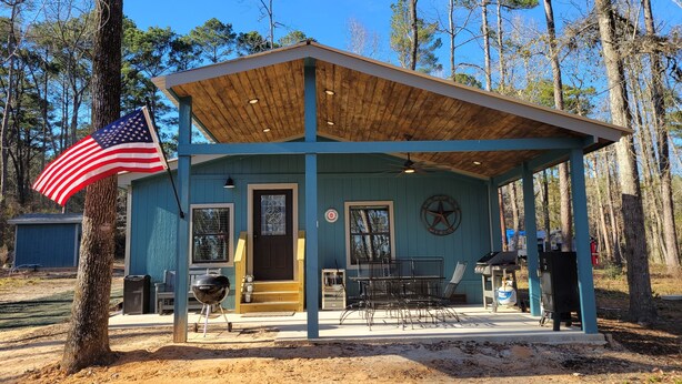 A small, single-story building with a blue exterior, a covered porch, a flag, a grill, and outdoor seating. - Cabin on a Secluded Cove -- READ DESCRIPTION REGARDING SAM RAYBURN WATER LEVEL (Broaddus)