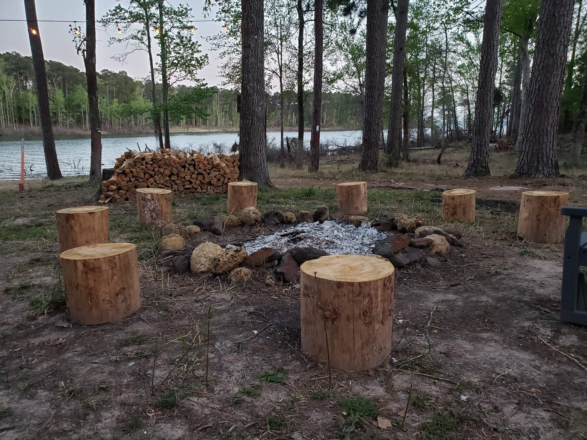 Una fogata rodeada de troncos de madera, con un montón de leña y un lago al fondo.