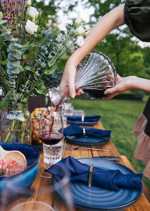Una persona sirve vino tinto en una copa en una mesa de comedor al aire libre, con un florero de flores y un bol de comida.