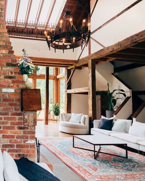 Ein geräumiger Wohnbereich mit hoher Decke, freiliegenden Holzbalken, einer Chandelüster, einem weißen Sofa, einem gemusterten Teppich und einer Ziegelwand.