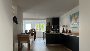 A modern kitchen with a central island, wooden dining table, and chairs. There is a view of greenery outside through large windows. - House fig trees Roch Priol (Quiberon)